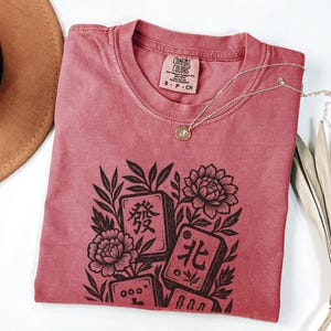 以下が含まれることがあります： 赤みがかったTシャツに、花と麻雀牌の黒いグラフィック。シャツはクルーネックで、首元に小さなタグが付いています。ペンダント付きのシルバーネックレスがシャツの上にかけられています。
