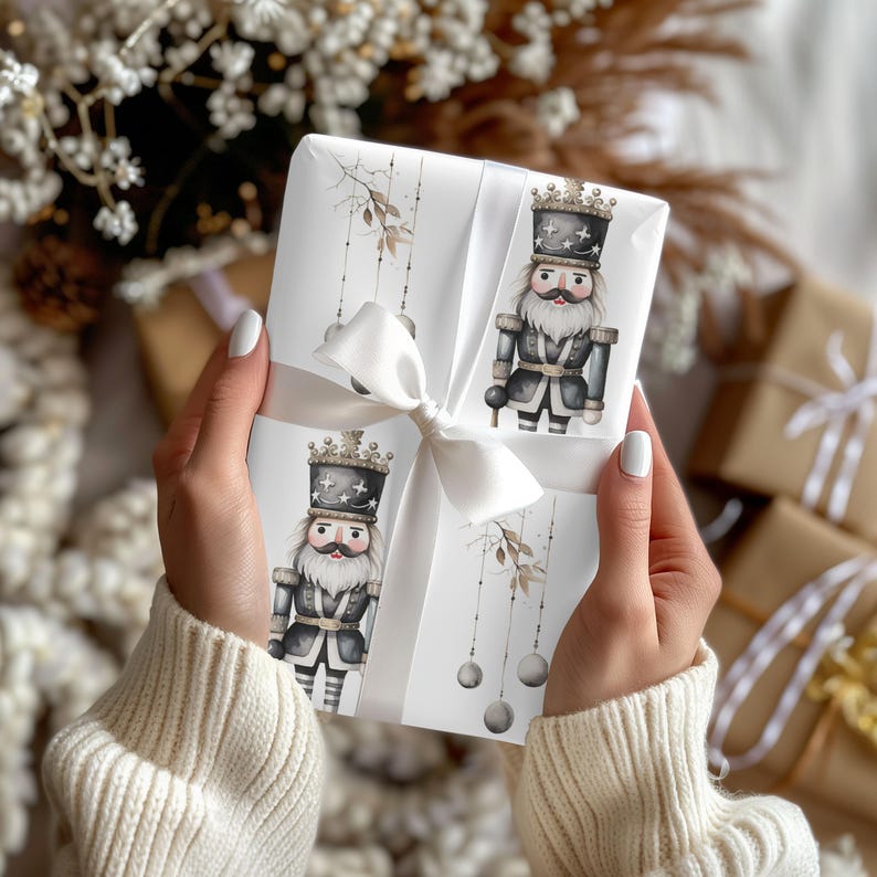 May include: A white gift box with illustrations of nutcrackers in black and grey uniforms with gold crowns. The box is tied with a white satin ribbon and bow. Other wrapped gifts are in the background.
