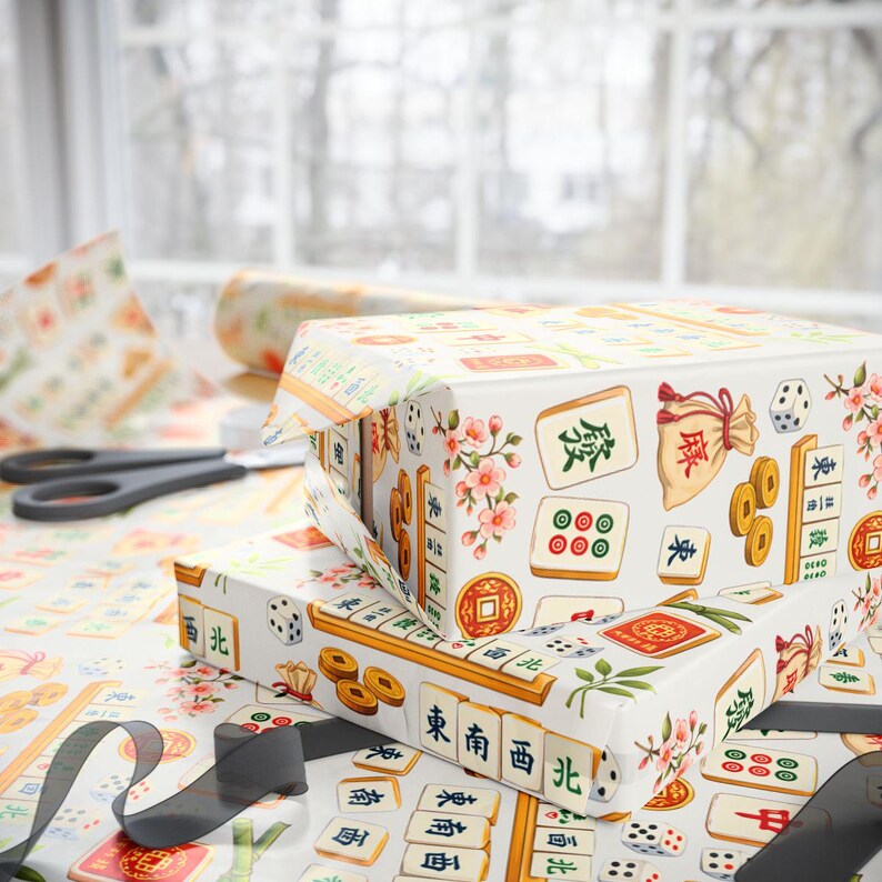 May include: Two gift boxes wrapped in patterned paper featuring Mahjong tiles, dice, coins, and floral accents. The boxes are on a table with matching wrapping paper, scissors, and a black ribbon. The design includes Chinese characters.