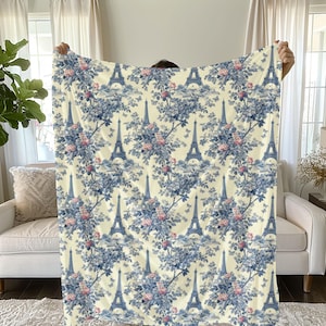 May include: A white and blue patterned throw blanket features a repeating design of the Eiffel Tower and floral bouquets. The background is a light cream color.