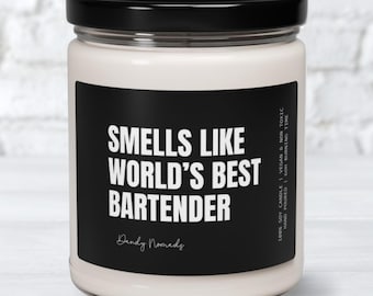 May include: A white candle with a black label that reads "Smells Like World's Best Bartender" with the text "Dandy Nomads" below. The candle is in a glass jar with a black lid.