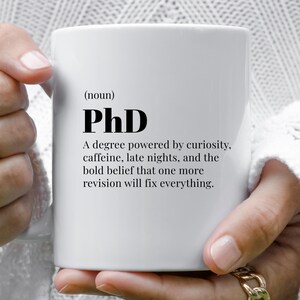 Peut inclure: Tasse en céramique blanche avec le texte "(nom) PhD" en noir. La description de la tasse comprend "Un diplôme alimenté par la curiosité, la caféine, les nuits tardives et la ferme conviction qu'une révision de plus corrigera tout."