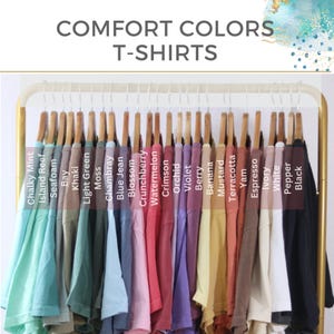 May include: A display of Comfort Colors T-shirts in various hues, including Chalky Mint, Seafoam, and Crimson. The shirts are neatly arranged on wooden hangers, showcasing a range of colors from light pastels to deep tones. The text "COMFORT COLORS T-SHIRTS" is at the top.