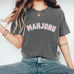 May include: Dark gray t-shirt with the word "MAHJONG" in pink letters with a green outline. The shirt is a casual style, suitable for everyday wear. The text is arched across the chest.