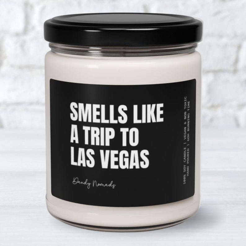 May include: A white soy candle with a black label that reads "Smells Like A Trip To Las Vegas" and "Dandy Nomads". The candle is in a glass jar with a black lid.