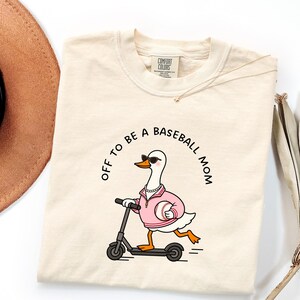 May include: A cream-coloured t-shirt with the text "OFF TO BE A BASEBALL MOM." The shirt features a cartoon duck wearing sunglasses and a pink jacket, riding a scooter and holding a baseball. The shirt has a Comfort Colors tag.