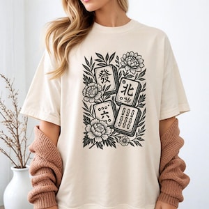 以下が含まれることがあります： クリーム色のTシャツ。白黒の麻雀牌と花のグラフィックが特徴。デザインには漢字と様式化された花が含まれており、ヴィンテージな美学を演出。ゆったりとしたフィット感のシャツです。