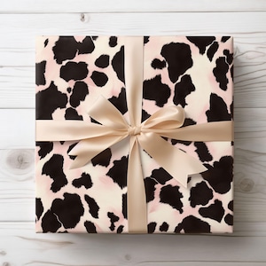 Puede incluir: Una caja de regalo envuelta en papel de regalo de estampado de vaca rosa y negro con una cinta de satén beige atada en un lazo.