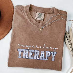 May include: A brown Comfort Colors t-shirt with the words "respiratory THERAPY" in white script and blue gingham block letters. A silver necklace with a small pendant rests on the shirt. A brown hat is partially visible in the upper left corner.