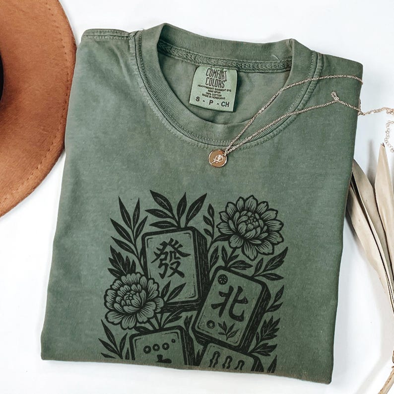 以下が含まれることがあります： オリーブグリーンのTシャツ。黒い麻雀牌と花のグラフィックがプリントされています。Tシャツはクルーネックで半袖です。ペンダント付きのゴールドネックレスがTシャツの上にかけられています。