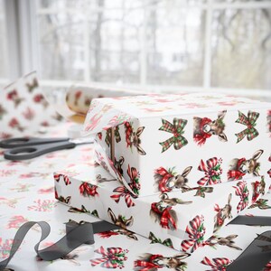 May include: Two wrapped gift boxes with white wrapping paper featuring a pattern of deer heads wearing red and green scarves and bows. A roll of matching wrapping paper and scissors are visible in the background, along with a roll of wrapping paper.