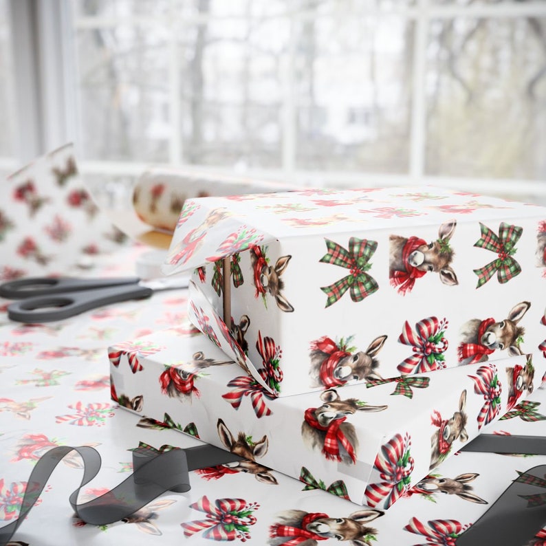 May include: Two wrapped gift boxes with white wrapping paper featuring a pattern of deer heads wearing red and green scarves and bows. A roll of matching wrapping paper and scissors are visible in the background, along with a roll of wrapping paper.