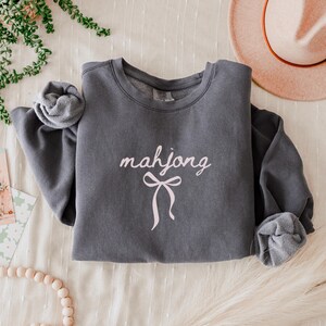 May include: A charcoal gray sweatshirt with the word "mahjong" in a cursive font and a pink bow graphic. The sleeves are cuffed. A pink hat is in the background.