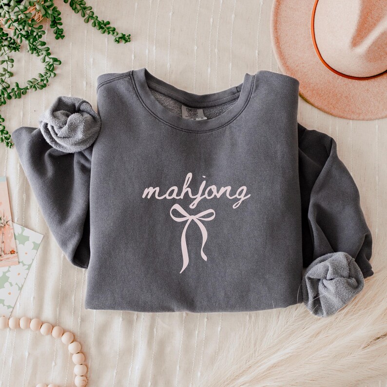 May include: A charcoal gray sweatshirt with the word "mahjong" in a cursive font and a pink bow graphic. The sleeves are cuffed. A pink hat is in the background.