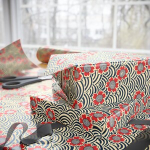 Puede incluir: Cajas de regalo envueltas en papel estampado con flores rojas y dise&ntilde;os de olas azul marino. Las cajas est&aacute;n apiladas sobre una mesa con tijeras y un rollo de papel de regalo. Una cinta negra est&aacute; en primer plano.