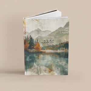 K&ouml;nnte beinhalten: Ein Hardcover-Journal mit einem Landschaftsdesign mit Bergen, einem See und B&auml;umen. Der Einband hat einen Aquarell-Effekt mit dem Jahr "2026" in Schwarz gedruckt. Das Journal ist ge&ouml;ffnet und zeigt wei&szlig;e Seiten.