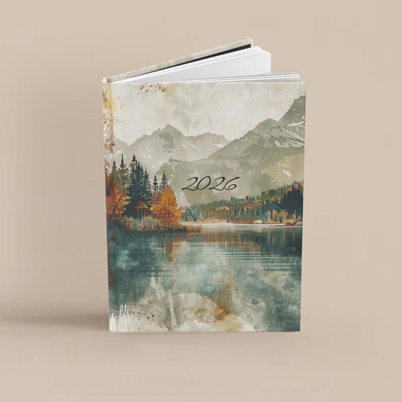 K&ouml;nnte beinhalten: Ein Hardcover-Journal mit einem Landschaftsdesign mit Bergen, einem See und B&auml;umen. Der Einband hat einen Aquarell-Effekt mit dem Jahr "2026" in Schwarz gedruckt. Das Journal ist ge&ouml;ffnet und zeigt wei&szlig;e Seiten.