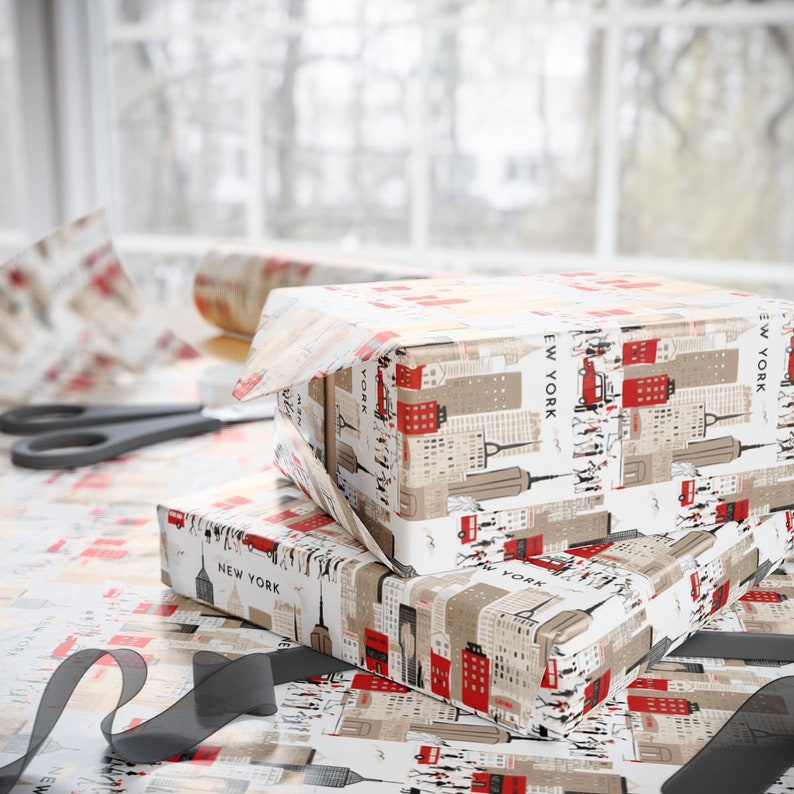 May include: Two gift boxes wrapped in white and beige wrapping paper with a red and black New York City skyline design. The boxes are sitting on a table covered in the same wrapping paper.
