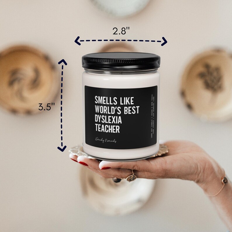 May include: A white candle with a black lid and a black label that reads "Smells Like World's Best Dyslexia Teacher." The candle is 3.5 inches tall and 2.8 inches wide. The candle is held in a person's hand.