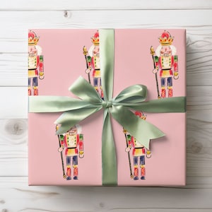 May include: A wrapped gift with a light pink wrapping paper featuring a repeating pattern of watercolor nutcracker soldiers. A sage green satin ribbon is tied in a bow around the gift. The background is a white wooden surface.