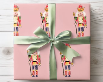 May include: A wrapped gift with light pink wrapping paper patterned with watercolour nutcracker soldiers. A sage green satin ribbon is tied in a bow. The background is a white wooden surface.