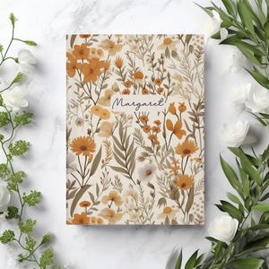 May include: A notebook featuring a floral design with orange, yellow, and white flowers and green leaves on a cream background. The name "Margaret" is written in cursive on the cover.
