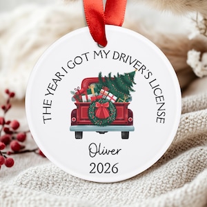 Pu&ograve; includere: Un ornamento rotondo in ceramica bianca con un nastro rosso. Il design raffigura un pickup rosso che trasporta un albero di Natale e regali, con una ghirlanda sul portellone. Il testo recita: THE YEAR I GOT MY DRIVER'S LICENSE, Oliver, 2026.