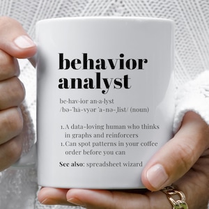 May include: White ceramic mug with the words "behavior analyst" in bold black font. The mug includes a definition, pronunciation, and two humorous statements about the profession. The mug is held by a person.