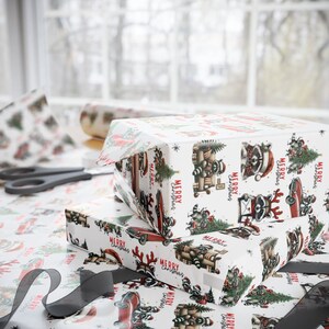 May include: Two wrapped gifts with white wrapping paper featuring a repeating pattern of raccoons, Christmas trees, and red cars. The words "MERRY CHRISTMAS" are printed on the paper. A pair of scissors and a roll of wrapping paper are in the background.