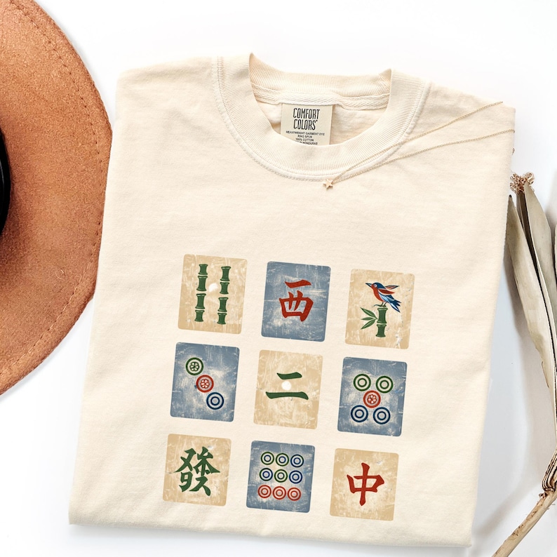 May include: Cream-colored t-shirt featuring a colorful Mahjong tile design. The graphic includes various tiles with bamboo, circles, and Chinese characters. The shirt has a tag that reads "Comfort Colors".