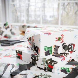 Puede incluir: Dos cajas de regalo envueltas con un fondo blanco, con un patr&oacute;n repetido de gatos negros con bufandas y gorros de Pap&aacute; Noel. Las cajas est&aacute;n sobre una mesa con papel de regalo, tijeras y una cinta negra.