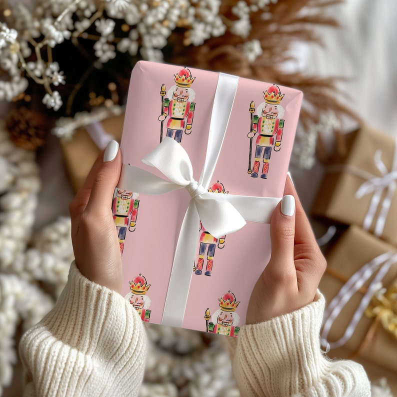 May include: A wrapped gift with pink wrapping paper featuring a repeating pattern of watercolor nutcracker soldiers. The gift is tied with a white satin ribbon and bow. The background includes other wrapped gifts and decorative elements.
