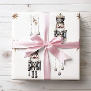 May include: A wrapped gift with white paper featuring illustrations of nutcrackers in black and grey uniforms. The gift is tied with a light pink satin ribbon and bow. Gold star and ornament details complete the festive design.