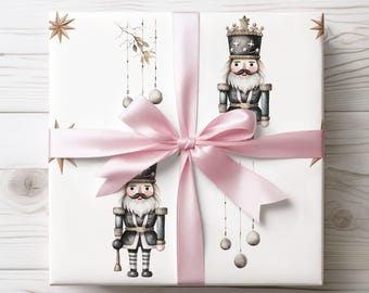 May include: A wrapped gift with white paper featuring illustrations of nutcrackers in black and gray uniforms. The gift is tied with a light pink satin ribbon and bow. Gold star and ornament details complete the festive design.