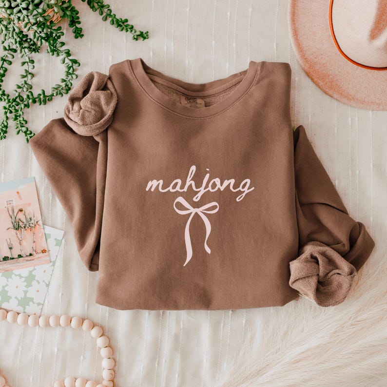 May include: A brown sweatshirt with the word "mahjong" in a cursive font and a bow graphic in light pink. The sweatshirt has long sleeves and a crew neck. The sleeves have a gathered detail at the shoulders.