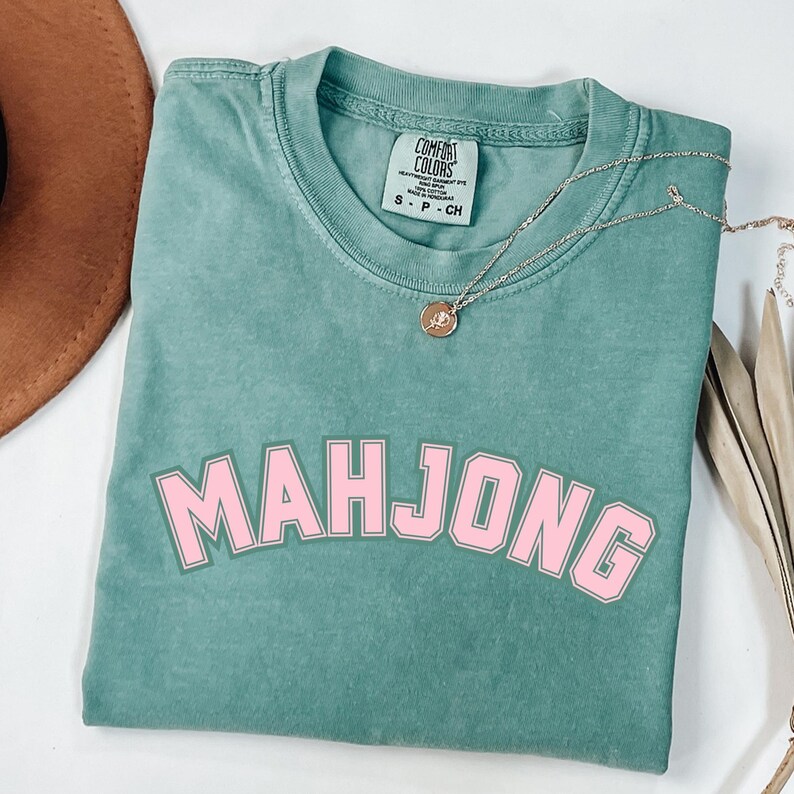 May include: A teal Comfort Colors t-shirt with the word "MAHJONG" in pink block letters. A gold necklace with a pendant rests on the shirt. The shirt is folded, and a brown hat is partially visible in the background.