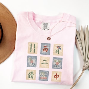 May include: A light pink t-shirt with a graphic of nine Mahjong tiles. The tiles are in various colors and feature bamboo, circles, and Chinese characters. A gold necklace with a pendant is on the shirt.