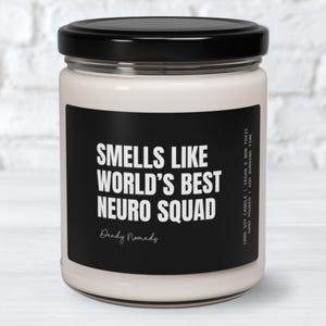 K&ouml;nnte beinhalten: Eine wei&szlig;e Kerze mit einem schwarzen Etikett, auf dem "Smells like World's Best Neuro Squad" steht, darunter der Text "Dandy Nomads". Die Kerze befindet sich in einem Glasgef&auml;&szlig; mit einem schwarzen Deckel.