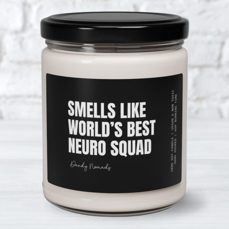 K&ouml;nnte beinhalten: Eine wei&szlig;e Kerze mit einem schwarzen Etikett, auf dem "Smells like World's Best Neuro Squad" steht, darunter der Text "Dandy Nomads". Die Kerze befindet sich in einem Glasgef&auml;&szlig; mit einem schwarzen Deckel.