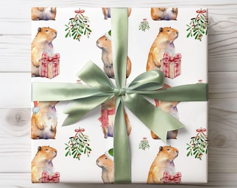 Pode incluir: Papel de embrulho branco com um padrão repetido de capivaras em aquarela com presentes, azevinho e acessórios festivos. Uma fita e um laço de cetim verde sálvia envolvem o presente. O fundo é um padrão de grão de madeira clara.