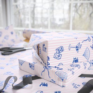 May include: A wrapped gift box with pink and white striped wrapping paper. The paper is decorated with blue illustrations of fans, dice, and other objects. A pair of scissors and a roll of matching paper are in the background.