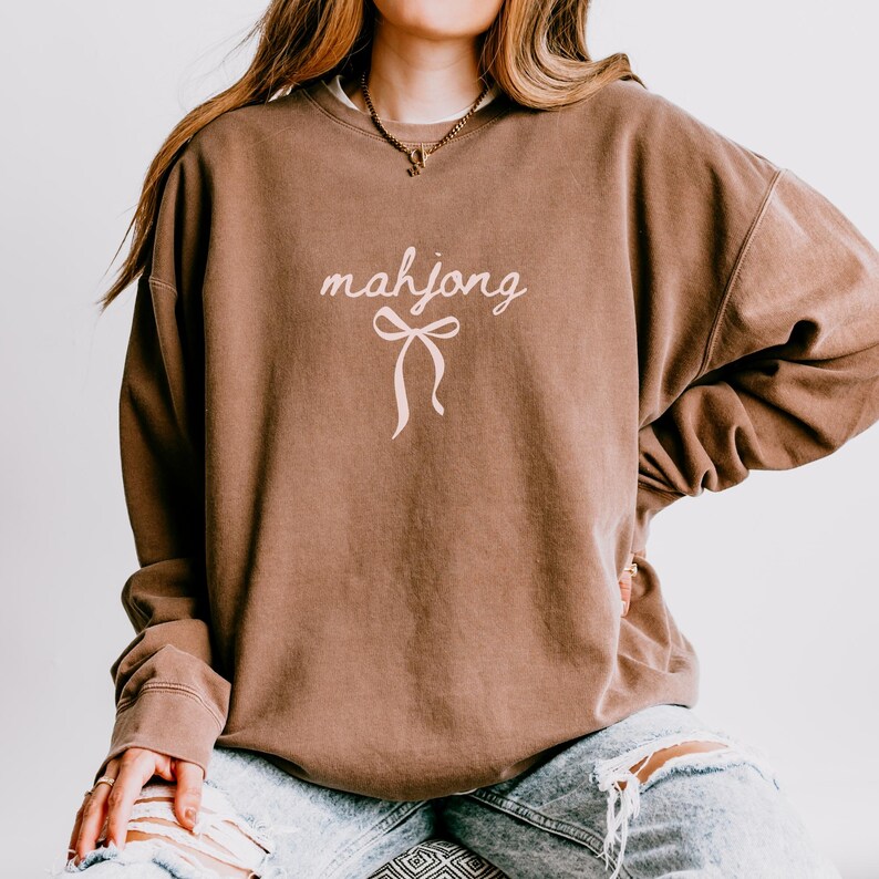 May include: A brown sweatshirt with the word "mahjong" in a cursive font and a bow graphic. The sweatshirt has long sleeves and a relaxed fit. The garment is a solid brown color.