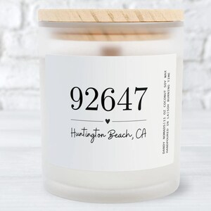 Puede incluir: Una vela blanca con una tapa de madera y una etiqueta que dice "92647 Huntington Beach, CA". La etiqueta también incluye el texto "DANDY NOMADS 11 OZ COCONUT SOY WAX HANDPOURED IN LA 50H BURNING TIME".