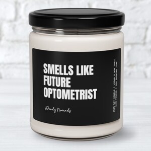 May include: A white candle with a black label that reads "Smells Like Future Optometrist" and "Dandy Nomads". The label also includes the details "100% Soy Candle | Vegan & Non Toxic | Hand Poured | 60h Burning Time".