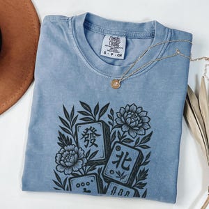 以下が含まれることがあります： マージャン牌と花の黒いグラフィックが特徴の、ライトブルーのComfort Colors Tシャツ。クルーネックと半袖です。ペンダント付きのゴールドネックレスがシャツの上にかけられています。