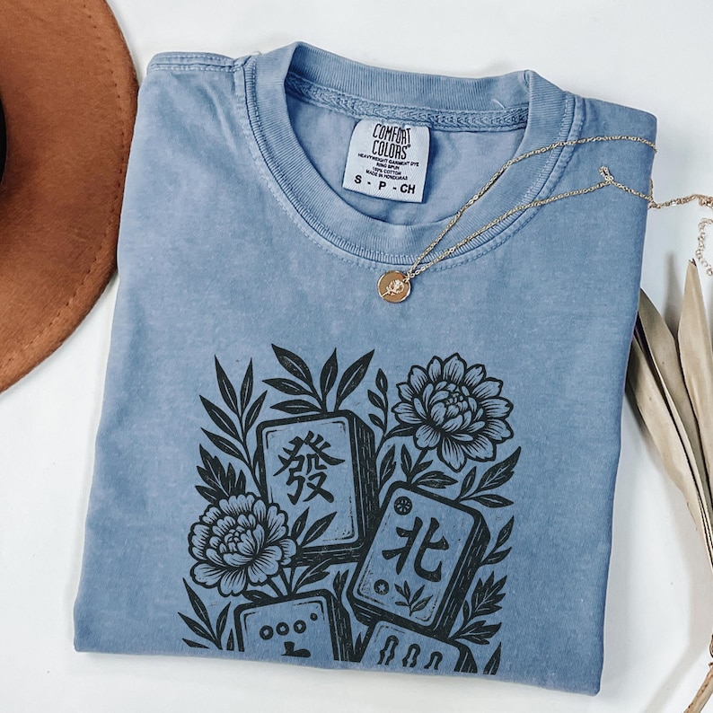 以下が含まれることがあります： マージャン牌と花の黒いグラフィックが特徴の、ライトブルーのComfort Colors Tシャツ。クルーネックと半袖です。ペンダント付きのゴールドネックレスがシャツの上にかけられています。