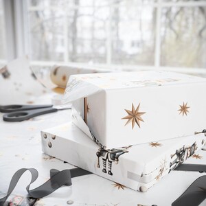 May include: Two white gift boxes wrapped in patterned paper with gold star and robot illustrations. The boxes are stacked on a table with scissors and a roll of wrapping paper. A black ribbon is in the foreground.