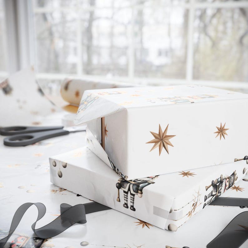 May include: Two white gift boxes wrapped in patterned paper with gold star and robot illustrations. The boxes are stacked on a table with scissors and a roll of wrapping paper. A black ribbon is in the foreground.