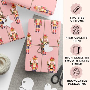May include: Pink wrapping paper with a repeating pattern of watercolor nutcrackers in red, yellow, and blue. The wrapped gifts are tied with twine and adorned with white heart-shaped tags. The image also shows text that says "TWO SIZE OPTIONS", "HIGH-QUALITY PRINT", "HIGH GLOSS OR SMOOTH MATTE FINISH", and "RECYCLABLE PACKAGING".