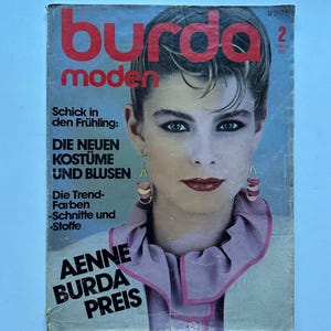 Op de afbeelding: Een vintage Burda Moden tijdschrift uit februari 1983 met op de cover een portret van een persoon met kort haar, gekleed in een lichtgekleurd kledingstuk met kraag, roze bies en gelaagde halvemaanvormige oorbellen. Tekst bevat Burda moden.